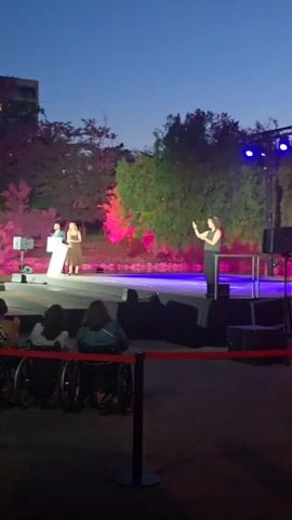 The video shows an outdoor evening event on a stage. There are people in wheelchairs in the foreground, with a red rope barrier. On the stage, a man in a suit stands at a podium. A woman in a brown dress stands next to him. Another woman in black pants and a black top is dancing. The stage is lit with pink and blue lights. Trees and a building are visible in the background. The sky is dark blue, indicating it is dusk or nighttime.