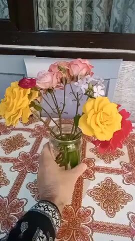 A hand holding a glass jar with flowers is seen in the center of the frame. The jar contains a bouquet of roses in various colors including yellow, pink, red, and some smaller white flowers. The flowers are arranged in a cylindrical glass jar filled with water. The hand appears to be gently adjusting the position of the jar. The background shows a patterned tablecloth and a window with curtains.