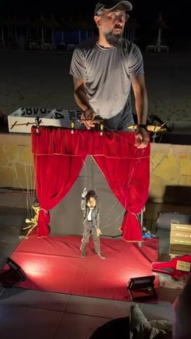 A man in a gray t-shirt and glasses operates a marionette from behind a red velvet curtain. The marionette, dressed in a black and white suit and a black hat, is suspended by strings and appears to be dancing. The scene is set outdoors at night, with beach chairs visible in the background. The stage is illuminated by spotlights.