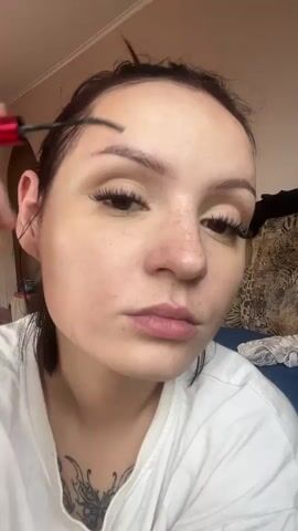 A close-up shot shows a young woman applying makeup to her eyebrows. She is holding a red and black eyebrow brush to her left eyebrow. Her hair is dark and pulled back. She is wearing a white t-shirt. Her face is mostly clear, with some freckles across her nose and cheeks. She has long, dark eyelashes. The background is blurred, but appears to be a room with patterned wallpaper and furniture.