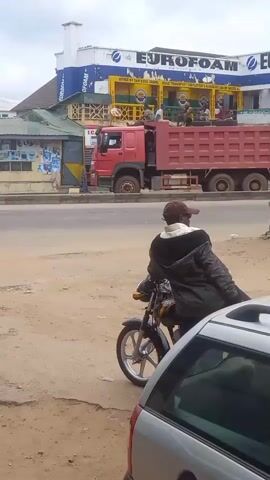 The video shows a street scene in an urban environment, likely in Sardauna, Nigeria, under an overcast sky. In the foreground, a man wearing a dark jacket and a cap sits on a motorcycle, facing away from the camera. Part of a silver car is also visible in the bottom right corner.

Across the road, traffic flows in both directions. Initially, a red dump truck drives from left to right, followed by a silver sedan moving from right to left. Subsequently, a large yellow dump truck, with "SINOTRUK" (partially visible as "SINDYRUH") branding on its side, drives from left to right. A blue car is also parked further down the road on the right side.

On the far side of the road, a multi-story commercial building is prominent. Its upper facade displays large "EUROFOAM" signs in blue and white. Below these, there are other signs, including "OFFICE OF YAN DOYA CHAPEL ROAD TRANSPORT EMPLOYERS ASSOCIATION OF NIGERIA" and "I.C. MARC INT'L CO." Solar panels are displayed outside the "I.C. MARC INT'L CO." storefront. The scene depicts typical daytime activity on a busy road.