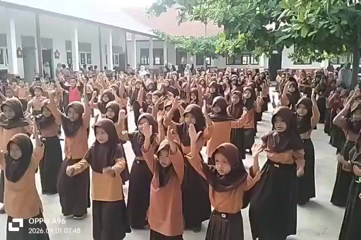 A large group of young girls, dressed in school uniforms consisting of brown hijabs and skirts, and tan tops, are gathered in an outdoor courtyard. The girls are arranged in rows, and most appear to be in the process of performing a synchronized dance or exercise routine. They are clapping their hands and moving their arms in unison. Some girls are closer to the camera, while others are further back. The courtyard is paved and appears to be part of a school campus, with buildings visible in the background and trees providing shade. The lighting suggests it is daytime.