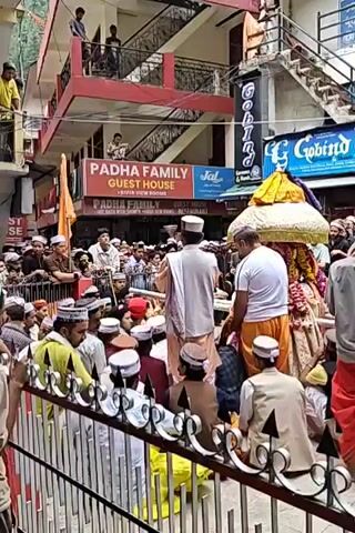 This video shows a bustling street scene in Kasol, India, during what appears to be a local procession or festival. A large crowd of people, predominantly men, are gathered on the street. Many of the men are wearing distinctive caps, possibly traditional attire for the region.

At the center of the crowd, a decorated structure, possibly a palanquin or a small chariot adorned with flowers and colorful fabrics, is being carried or moved by several individuals. Some men in white and orange garments are actively involved with this structure. An orange flag is also visible within the crowd, near the decorated object.

The street is lined with multi-story buildings, some with balconies where other people are observing the event from above. One prominent sign reads "PADHA FAMILY GUEST HOUSE" with "RIVER VIEW ROOMS" visible below it. Another sign for "GOVIND" is partially visible on an adjacent building. The weather appears to be clear and bright, indicating daytime. The atmosphere suggests a cultural or religious event, with many participants and onlookers.