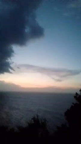 The video shows a wide shot of the ocean and sky at what appears to be dusk or dawn. Dark clouds are visible in the upper left of the frame, with lighter clouds stretching across the horizon. The ocean is dark and appears to have waves. In the bottom right corner, a dark silhouette of trees is visible. In the distance along the coastline, lights from a town or city are visible. The video is shaky, suggesting it was hand-held.
