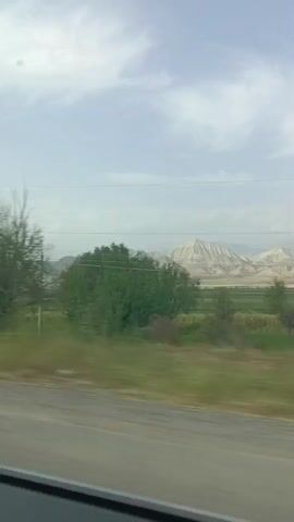 The video shows a moving view from a vehicle traveling on a road. The landscape outside consists of fields, trees, and mountains in the distance. Power lines and utility poles are visible throughout the video, indicating the presence of infrastructure. There are also a few small buildings or structures along the roadside. The weather appears to be overcast with some clouds in the sky. The video seems to capture a rural or semi-rural environment.