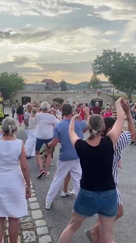 A group of people are dancing outdoors during the day. The sky is cloudy with the sun peeking through. The ground is paved with cobblestones. In the background, there are buildings, trees, and parked cars. Some people are dancing in pairs, while others are dancing in a line. There is a person standing to the left wearing a yellow shirt and a hat, possibly an instructor or DJ. The people are dressed in casual summer clothing. The atmosphere appears to be festive and lively.