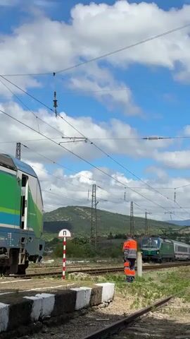 Two trains are visible on parallel tracks under a cloudy blue sky. On the left, a portion of a blue and green train is stationary, with its doors open. On the right, a green and white train is moving forward. A person in an orange uniform stands between the tracks, facing the moving train and appearing to be signaling. In the background, hills are covered in green foliage, and overhead electrical wires for the trains crisscross the sky. A red and white striped pole with a circular sign is also visible near the stationary train.
