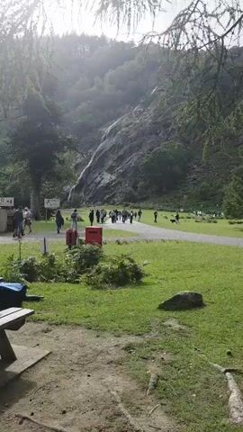 The video shows a view of a valley with a waterfall cascading down a rocky mountainside. In the foreground, there is a picnic table with some bags and water bottles on it. A path runs through a grassy area where several people are walking. Some are in a group, while others are walking individually or in smaller groups. There are also trash cans and signs visible along the path. The overall atmosphere appears to be a park or natural attraction. The lighting suggests it may be overcast or late in the day, with sunlight filtering through trees.