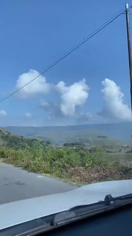 The video shows a point-of-view shot from inside a vehicle, looking out at a scenic landscape. A road curves to the left in the foreground, bordered by lush green vegetation and trees. Beyond the roadside greenery, rolling hills and mountains stretch into the distance under a clear blue sky dotted with fluffy white clouds. Power lines are visible crossing the sky. The dashboard and windshield wipers of the vehicle are visible at the bottom of the frame. The lighting suggests it is daytime, likely sunny. There are no people or animals clearly visible in the frame.