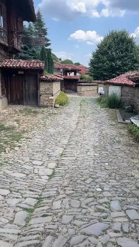 A cobblestone pathway winds through an old village. On the left, a two-story building with weathered wooden walls and stone foundations stands. Its exterior features multiple windows and a balcony on the upper level. A wooden door is visible on the ground floor, set within a stone wall with a tiled roof. Further down the pathway, similar stone-walled structures with wooden doors and tiled roofs line the street. Trees and lush greenery are visible in the background, and a cloudy blue sky stretches overhead. The scene is bathed in natural light, suggesting it was filmed during the daytime.