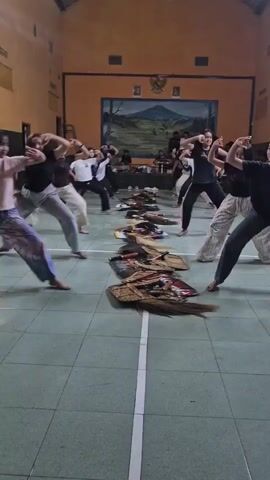A group of young women, likely students, are practicing a choreographed dance or martial art in what appears to be a gymnasium or large hall. They are dressed in casual clothing, primarily t-shirts and loose-fitting pants. The floor is tiled and marked with white lines. In the background, there is a mural depicting a landscape with mountains, and various figures are seated or standing near a table. Laid out on the floor in neat rows are what appear to be traditional Indonesian costumes or props, possibly related to the dance being practiced. The women are all in motion, striking similar poses with their arms raised and bodies bent, indicating a synchronized routine. The lighting is artificial and somewhat dim.