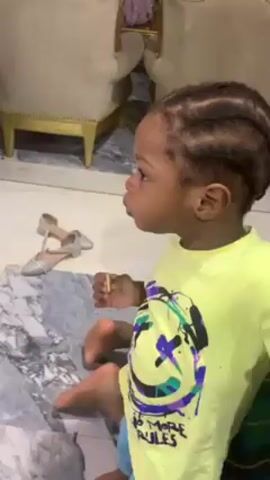 A young child stands barefoot on a marble floor, holding a small, crumbly biscuit. The child has braided hair and wears a bright yellow t-shirt with a smiley face design. They look directly at the camera. A pair of silver shoes sits on the floor behind them.  A beige chair with gold trim is partially visible in the background. The child is asked for a biscuit and holds it out slightly.