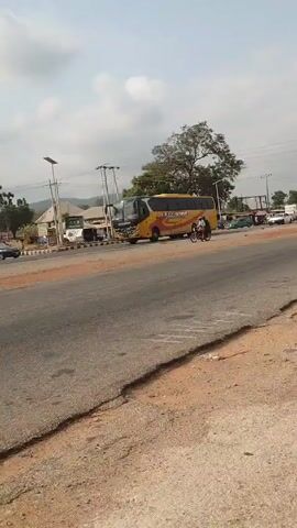 The video shows a road with a yellow bus stopped on the side.  A motorcycle drives down the road. Other cars are parked on the other side of the road.  There is a tree to the right, and some buildings and power lines visible in the background.  The road is cracked and the shoulder is sandy. The sky is cloudy.