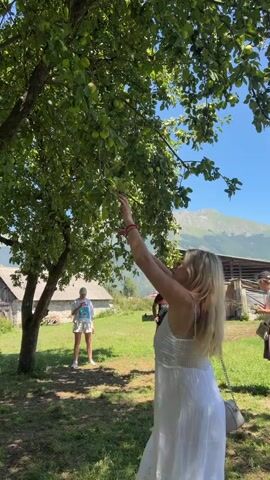 A blonde woman in a white dress reaches up to pick apples from a tree. Another person is visible in the background. The scene is outdoors with green grass, trees, and mountains in the distance under a blue sky. The woman then brings an apple to her mouth.