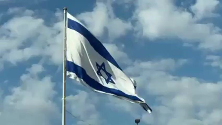 The video shows the flag of Israel waving in the wind against a blue sky with white clouds. The flag is white with two horizontal blue stripes and a blue Star of David in the center. The flag is attached to a metal pole.