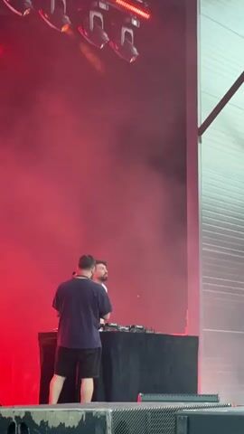 The video shows two DJs on a stage with red lighting and smoke. The DJ on the left is wearing a dark t-shirt and shorts, while the DJ on the right is wearing a white shirt. They are both standing behind a table with DJ equipment. In the background, there is a large brick wall with graffiti on it and a yellow sign with black text. A crowd of people is visible in front of the stage, many of them holding up their phones. The overall atmosphere appears to be that of an outdoor concert or festival.