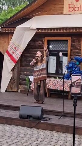 This video captures a performance outdoors on a wooden stage. A person stands center stage, singing into a microphone and gesturing with their raised hand. They are dressed in a patterned sweater and loose-fitting pants, giving them a bohemian or folk appearance. Behind the performer, a rustic wooden building with a prominent window serves as the backdrop. A large white banner with red and white embroidered patterns hangs from the left side of the stage, partially obscuring the building's facade.

The stage is set up for a performance, with a speaker on the left and a microphone stand on the right. A table draped with a patterned cloth is visible behind the performer, adorned with blue decorative elements that resemble large flowers. A smartphone on a tripod, recording the event, is positioned on the right side of the stage.

The setting appears to be a village or cultural center, suggested by the traditional architecture of the building and the folk-style banner. The overall atmosphere seems lively and engaging, with the performer conveying a sense of passion and connection with their audience. The presence of the recording device indicates that the event is being documented. The lighting suggests it is daytime, likely late afternoon or early evening. The video also includes a glimpse of trees in the background, hinting at an outdoor, possibly natural, setting. The word "Быги" is visible on the building, which, combined with the context "Старые Быги, Russia," indicates the location.