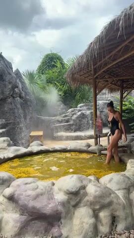 A young woman in a black swimsuit steps into a small, yellow pool of water filled with yellow flower petals. The pool is surrounded by gray rocks, with a bamboo shelter providing shade in the background. The surrounding area includes lush greenery, a small waterfall, and a cloudy sky. The woman moves into the pool and spreads her arms out to her sides, smiling at the camera.
