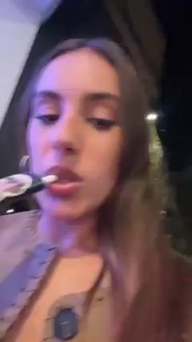 A woman with long brown hair is holding a black electronic cigarette with a picture of a dog wearing glasses on it to her lips. She takes a puff, then looks at the camera. She is wearing a beige jacket with dark stitching. The lighting is dim, and there are lights in the background.