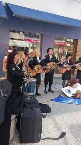A group of five people are performing music outdoors on a cobblestone street. Four of them are holding musical instruments and appear to be singing. Three of the performers are women, and two are men. The performers are dressed in black suits and white shirts, with some wearing hats. One performer is a woman of color wearing glasses. To the left of the group, there is a drum and several black bags, suggesting they are traveling musicians. In front of the performers, a blue tarp is spread on the ground with a stuffed monkey and a doll placed on top, possibly to collect tips. The background shows a building with large windows and awnings, and further back, outdoor seating areas of cafes and restaurants. The lighting suggests it is daytime.