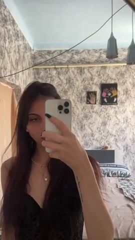A young woman with dark hair, wearing a black lacy top and leopard print pants, takes a selfie in a mirror. She has a piercing on her lip and is wearing a necklace. Her room is visible in the background, with wallpaper, a bed, and some artwork on the wall.