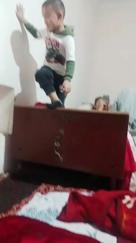A child stands on top of a dark wooden structure, possibly a bed headboard, with one hand against the white wall for balance. The child, dressed in a white hooded sweatshirt with green sleeves and dark pants, is facing the wall. In the background, a person lies on a bed using a phone, and another person is partially visible. The room has a patterned rug on the floor and the overall lighting is dim.