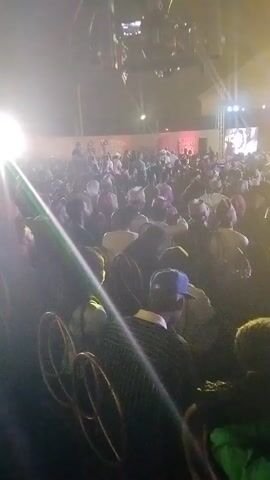 The video shows a large crowd of people gathered at an outdoor event, likely a concert or performance. A stage with a large screen displaying visuals is set up in the distance, illuminated by bright lights. The crowd is dense, with many people wearing traditional caps or hats. A complex lighting rig hangs above, casting beams of light across the scene. The overall atmosphere is lively, with the potential for social interaction among the attendees.