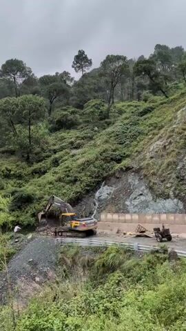 An excavator with the Volvo logo is on a road, digging into a hillside. The hillside is covered in trees and vegetation. There is a person standing next to the excavator. The road appears to be under construction or repair, with construction materials visible. The weather is overcast.