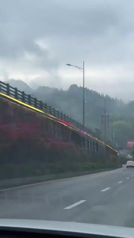 A car is driving on a wet road. There is a wall on the side of the road with colorful lights on it. In the background are mountains covered in mist.  There are lampposts on the side of the road with Chinese lanterns. Another car is in front. The sky is overcast.