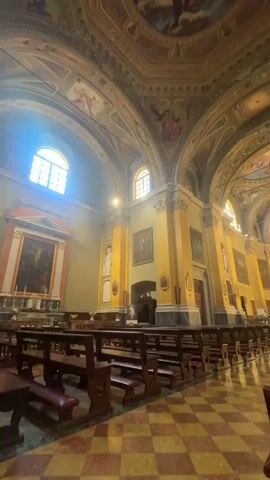 The video showcases the grand and ornate interior of a church in Binda, Italy, captured with a slow pan from left to right. The scene is dominated by high, arched ceilings adorned with intricate frescoes depicting religious figures and scenes, and a large central dome also painted with religious artwork.

Rows of dark wooden pews with red upholstered seats are arranged on a red and white checkered floor, leading towards the altar. Bright natural light streams in through tall arched windows on the sides of the church, illuminating the interior and creating sunbeams visible in the air. The walls are painted in warm yellow and green tones, featuring numerous religious paintings and statues set into alcoves or mounted on pillars.

A prominent pulpit with ornate carvings is visible on the right side of the nave. Towards the end of the video, a single person can be seen sitting quietly in one of the pews. The altar area at the front is brightly lit, adorned with candles and religious ornaments. The overall atmosphere is one of serene grandeur and historical richness. No visible text is clearly discernible in the video.