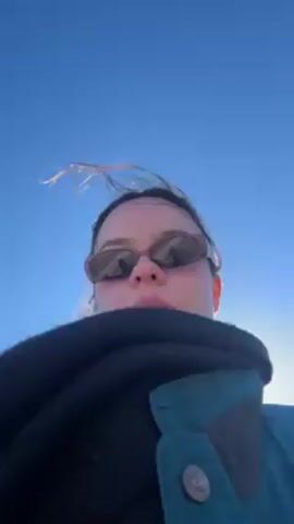 A person with sunglasses stands outside in bright sunlight under a clear blue sky.  The person is wearing a dark jacket with the collar pulled up. Their hair is blowing in the wind.