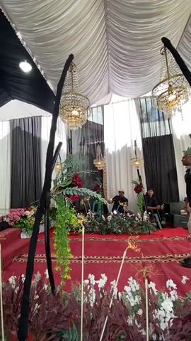 The low-angle shot captures a wedding reception, primarily focusing on the event's decorations and the stage where the bride stands with other guests. The tent is draped with white and black fabric. Two large chandeliers hang prominently, along with several smaller ones. The floor is covered with a red carpet. The stage is adorned with flowers in shades of red, pink, and white. There is also a lot of greenery, including hanging plants, which accentuates the decor. On the left, a woman in a light green outfit stands next to the bride, who is in a beige dress, with another woman in a purple dress next to her. On the right, three men are seated in a separate area of the stage, away from the women.