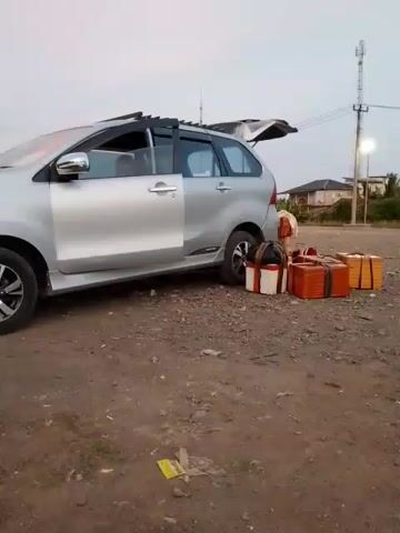 The video shows a silver minivan parked on a dirt surface with its rear hatch open. Several rectangular packages are placed on the ground next to the rear of the vehicle. The packages appear to be wrapped in brown and white materials. A person wearing an orange shirt and dark pants is partially visible in the driver's seat. The sky is overcast, suggesting it may be dusk or dawn. A utility pole with lights and a tower can be seen in the background, along with a building and some trees. The overall scene suggests someone is loading or unloading luggage from the car.