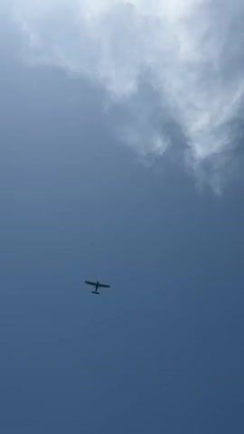 A small propeller airplane flies across a cloudy blue sky. The plane is silhouetted against the bright sky. The camera is pointed upwards at the plane and the sky.