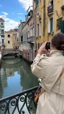 A woman with brown hair in a low bun, wearing a beige trench coat and a brown and tan handbag, stands on a bridge in Venice, Italy. She is holding a black smartphone horizontally and taking a photo of a canal. In the canal, there is a gondola with people in it, traveling under a small stone bridge. Buildings line both sides of the canal, with varying heights and colors, including yellow, gray, and red brick. The sky is blue with scattered white clouds. The bridge she is standing on has a black ornate railing.