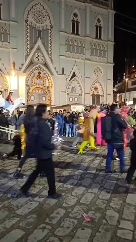 The video shows a band marching on a cobblestone street at night in front of a church with ornate Gothic architecture. The band consists of people in black suits playing trombone, cymbals, and other instruments. Several people are watching the band as it passes. There is a costume of a figure riding a white horse and other colorful costumes in the crowd. A fountain is visible in the background.