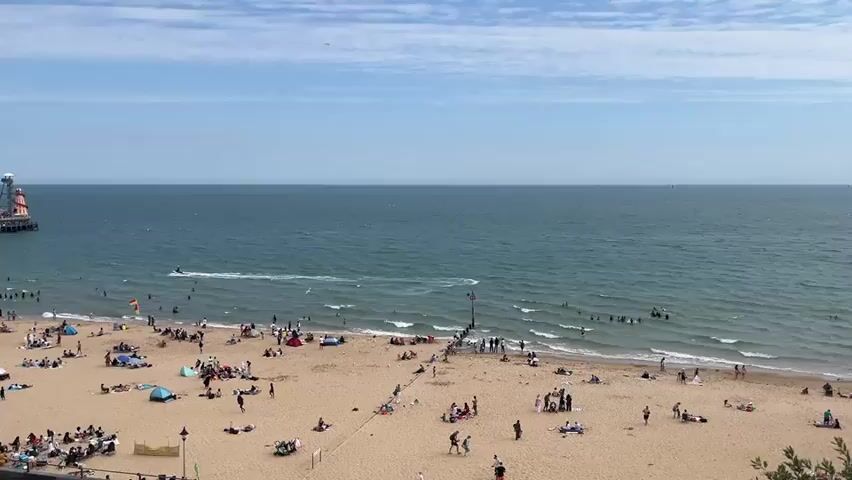 A wide shot captures a bustling beach scene on a sunny day. The sandy beach is populated with numerous people relaxing, sunbathing, and enjoying the water. Beach umbrellas and tents dot the sand, adding pops of color. In the background, a long pier extends into the calm, blue ocean. Several individuals are swimming or wading in the sea, while a jet ski creates a wake as it moves across the water. The sky above is a clear blue with scattered white clouds. The overall atmosphere is one of leisure and summer enjoyment.