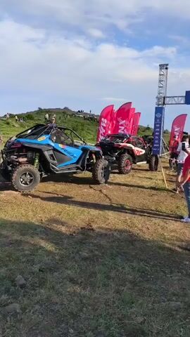 At 00:00, a blue and black off-road vehicle is parked on a dirt surface, facing left. Behind it, a red and white off-road vehicle is also parked. A large blue banner with the word "START" in white lettering is visible in the background, suspended from a metal structure. Several pink flags with black lettering are planted in the ground to the left of the blue vehicle. Two people are standing near the start banner, one wearing a white t-shirt and the other a red t-shirt. The ground is covered in dirt and grass. The sky is cloudy.

At 00:01, the blue and black off-road vehicle is still parked. The person in the red t-shirt is now facing towards the off-road vehicles. The start banner and flags remain in place.

At 00:02, the scene is largely unchanged. The off-road vehicles are stationary.

At 00:03, a person in a red shirt is walking away from the camera, towards the start line. The off-road vehicles are still parked.

At 00:04, the person in the red shirt has continued walking towards the start line and is now closer to the people near the banner. The off-road vehicles remain parked.