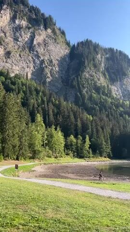 A scenic video pans across a lake nestled at the base of a steep, tree-covered mountain. The sky is a clear blue with a few wispy clouds. In the foreground, a grassy area borders a gravel path leading towards the lake. Two people are walking along the path near the water's edge. Further along the shore, a picnic table is occupied by three people, seemingly enjoying the view. The lake itself is calm and reflects the surrounding landscape. The overall impression is one of a peaceful and beautiful natural setting.