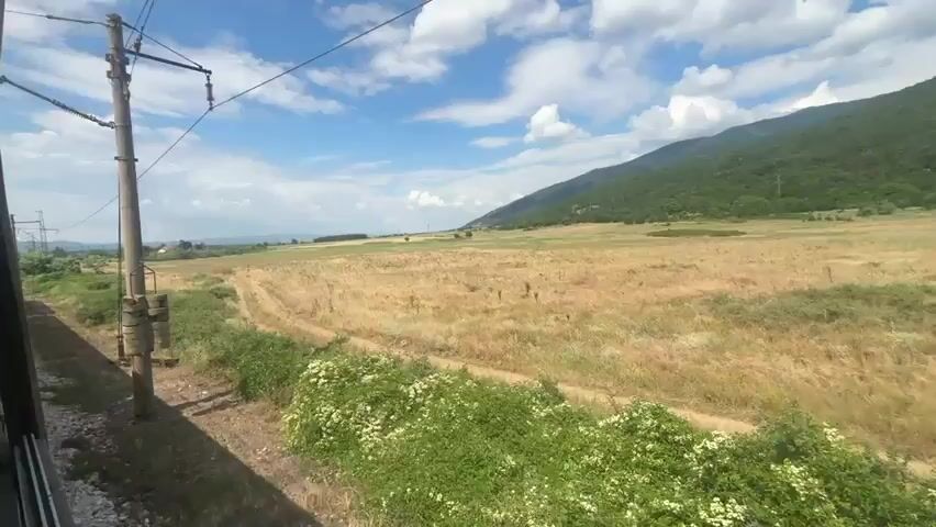 A train travels through a rural landscape on a sunny day. The view is from a window on the side of the train, showing a grassy field with dry yellow grass and scattered green bushes. In the distance, a line of green, tree-covered mountains stretches across the horizon under a bright blue sky with fluffy white clouds. A utility pole with wires is visible on the left side of the frame, and the side of the train car is visible on the far left. The train tracks are visible in the foreground.