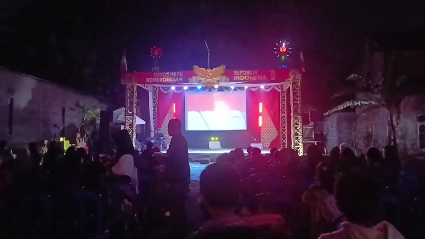 A night-time outdoor event is taking place with a stage set up at one end. The stage is adorned with red and white decorations, including the Indonesian flag. A large screen on the stage displays an image of the Indonesian flag. Two decorative fireworks-like lights are positioned on top of the stage, one on each side. In front of the stage, an audience is seated on chairs, facing the performance area. A few individuals are standing and walking around the periphery of the seating area. The environment appears to be a courtyard or open space surrounded by buildings, with trees visible in the background. The lighting is primarily from the stage, with some ambient darkness.