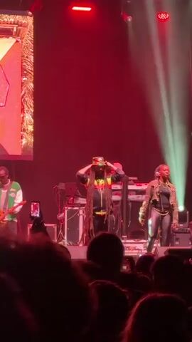 A live band performs on stage in front of a crowd.  The band includes a male vocalist with long dreadlocks and a bucket hat, a guitarist wearing a green and white jacket and sunglasses, a keyboardist, and a female singer.  Behind them, a screen shows an image of a man with the text "NINE MILE" displayed next to him. The stage is lit with red and white lights. The crowd is holding up their phones to record the performance.
