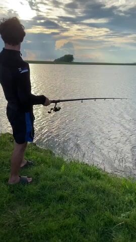 A young man with dark curly hair, wearing sunglasses, a black long-sleeved shirt, and black shorts with blue trim, stands on a grassy bank with his back to the camera, holding a fishing rod. He is looking out at a large body of water. The water is choppy with reflections of the sky. The sky is cloudy with sunlight breaking through. There is a distant tree on the opposite bank of the water. The man is wearing flip-flops.