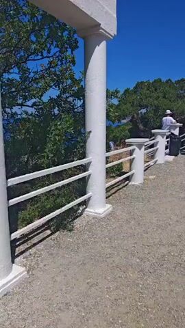 The video shows a group of people standing near a white railing on a gravel path. The railing is supported by white pillars. Behind the railing, there is a view of the sea and trees. Some people are looking out at the sea, while others are talking amongst themselves. The sky is blue and clear. A white building or structure is visible in the foreground.