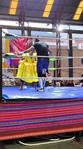 A man in a black shirt and blue wrestling pants faces a person in a full yellow dress inside a wrestling ring. The ring has a blue mat and is surrounded by white ropes. The man holds the person in the dress by the hand, helping them up. A referee stands nearby in a striped shirt. The ring is decorated with colorful banners and there is a crowd of people watching in the background. The building appears to be an indoor arena with a metal roof and support beams.