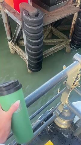 A person's hand holds a green tumbler in the foreground. The tumbler is green with a black lid. Behind the tumbler, a dock extends into green water. The dock is made of concrete and metal supports. Several black tires are stacked vertically on a metal pole, acting as a fender. Further in the background, a ferry is docked. The ferry is white with red accents and has a ramp leading onto it. The sky above is partly cloudy.
