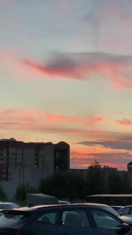 A vertical video shows a sunset over a city. The sky is filled with pink and orange clouds. In the foreground, there are several cars parked in what appears to be a parking lot. Behind the cars, there are buildings and trees. The lighting suggests it is dusk. The camera is mostly stationary, with slight movements. There are no people visible.
