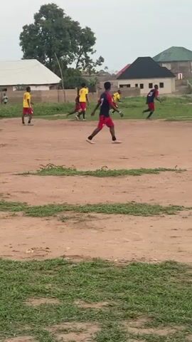 The video shows a group of people playing soccer on a dirt and grass field, with houses in the background. Most of the players are wearing red and blue uniforms, while others are wearing yellow jerseys. They are running around the field, presumably chasing the ball. One player in red and blue is walking. The weather appears overcast.