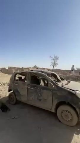 The video shows a desolate scene in what appears to be a war-torn or severely damaged village under a clear, bright blue sky. In the foreground, there is a badly damaged, muddy car with no roof, parked beside crumbling brick structures and piles of rubble. The car's interior is visible and stripped. Further in the background, there are more damaged buildings, some still standing but clearly in disrepair, with debris scattered around. Two men are visible, wearing traditional clothing, and appear to be examining the damage. Overall, the scene conveys a strong sense of destruction and desolation.