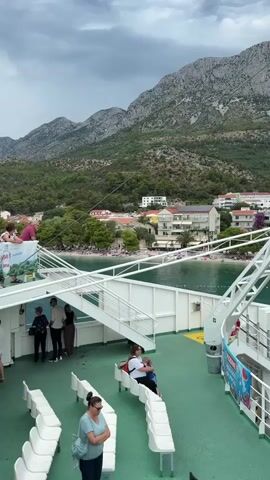The video shows a view from the deck of a ferry, with passengers seated on benches. In the foreground, several people are visible, some standing and some sitting. White benches are arranged in rows on the green deck. In the background, a body of water is visible, leading to a coastline with a beach crowded with people. Behind the beach, there are buildings, trees, and a large, mountainous landscape under a cloudy sky. The ferry appears to be moving or docked near the shore.